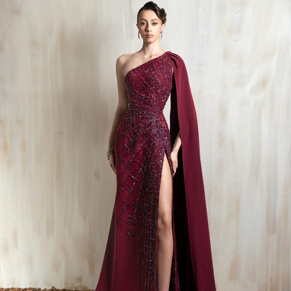 Burgundy one-shoulder gown with beaded detailing and a dramatic cape, designed for elegant occasions. Shop now at our exclusive clothing website.