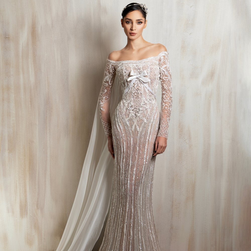 Ivory sheer fitted gown with off-shoulder lace, long sleeves, bow bodice & chiffon cape. Shop now at our exclusive clothing website.