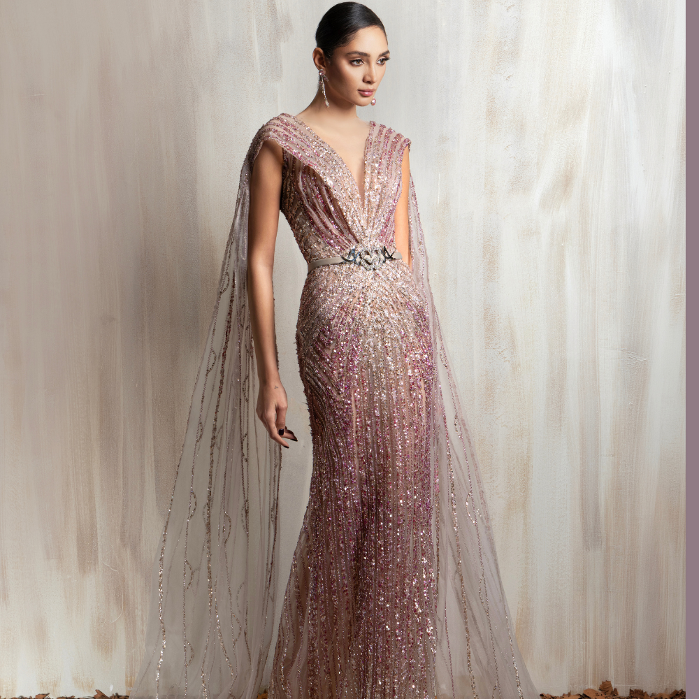 Rose gold beaded gown with a deep V neckline and flowing sheer cape, crafted for elegant occasions. Shop now at our exclusive clothing website.