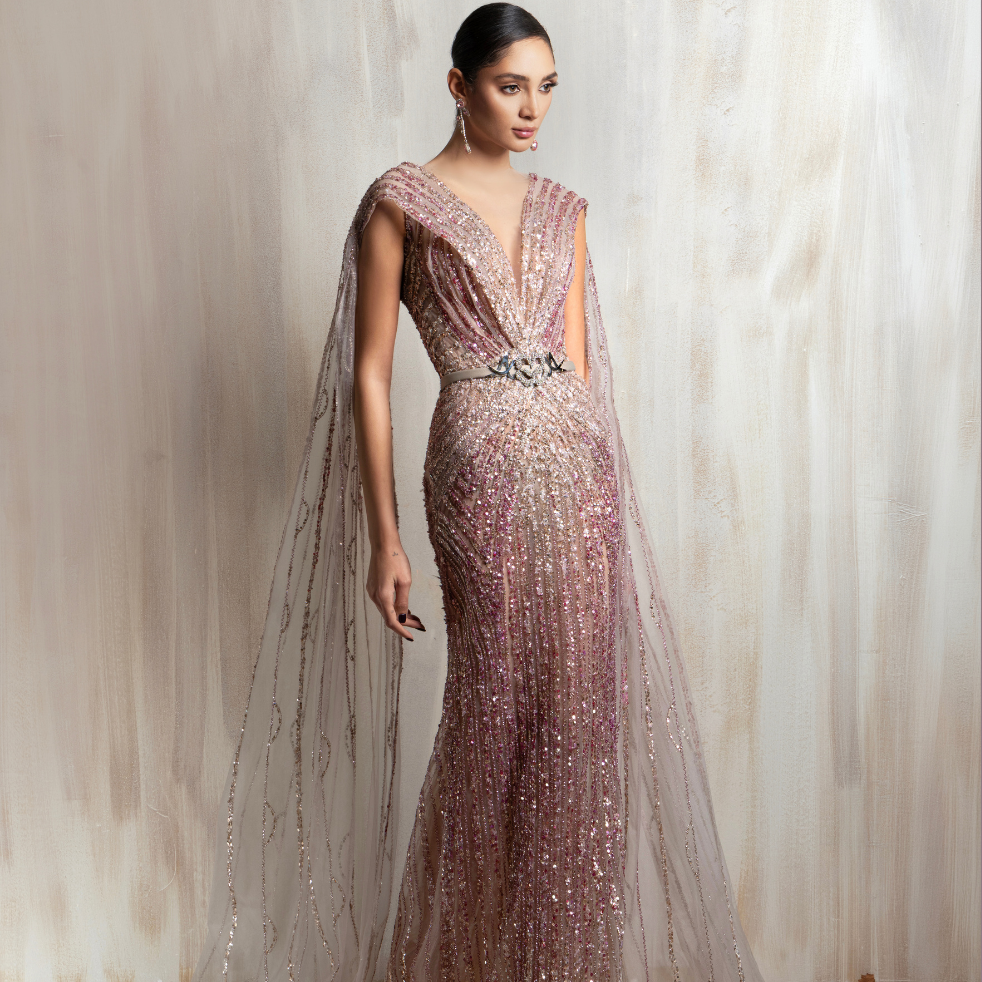 Rose gold beaded gown with a deep V neckline and flowing sheer cape, crafted for elegant occasions. Shop now at our exclusive clothing website.