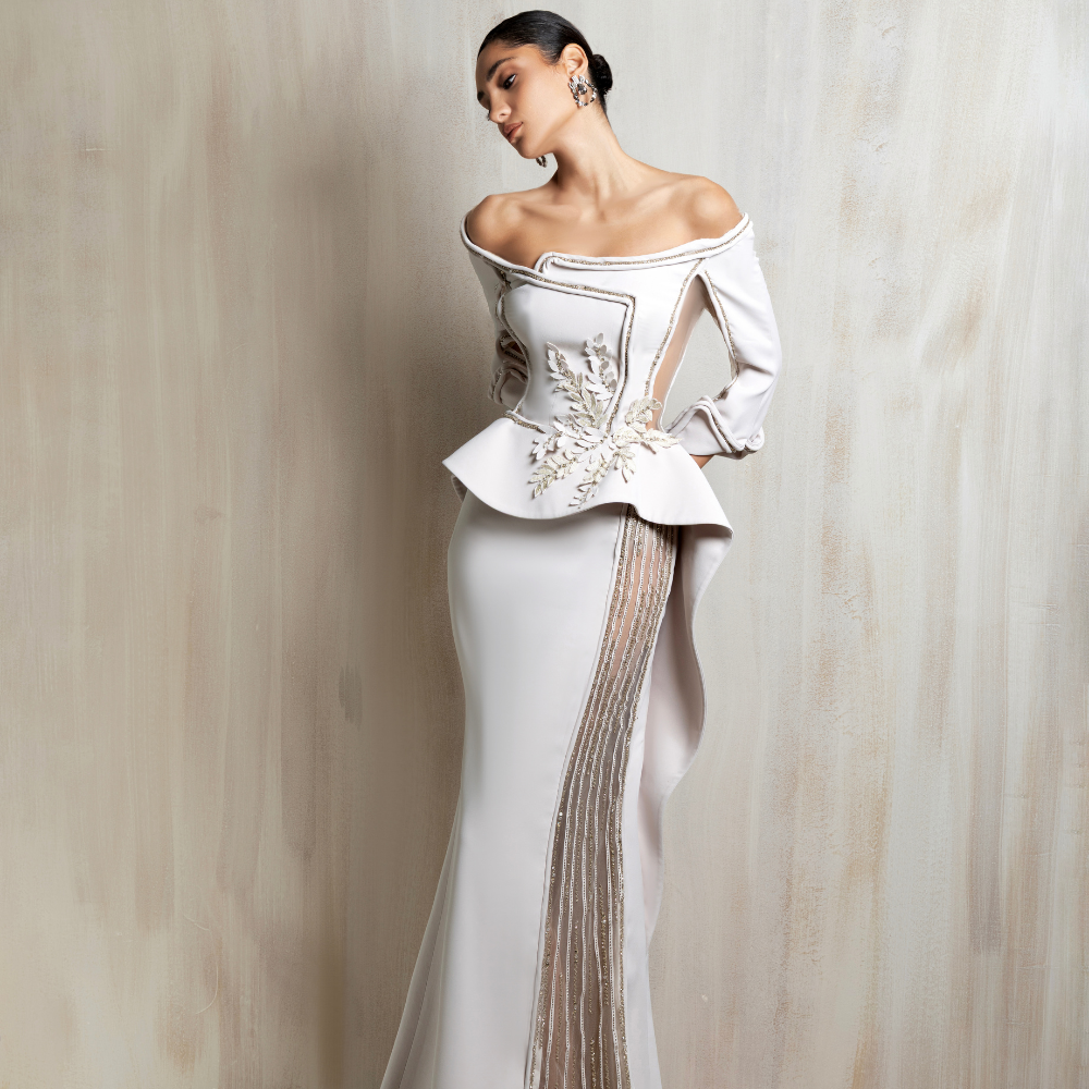 Off-shoulder off-white gown with a peplum bodice, floral appliqué waist, and sheer beaded panel. Shop now at our exclusive clothing website.