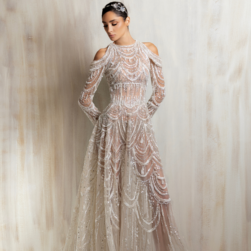 Sheer off-white gown with a high neckline, cold-shoulder sleeves, and cascading beadwork in layered draped patterns. Shop now at our exclusive clothing website.