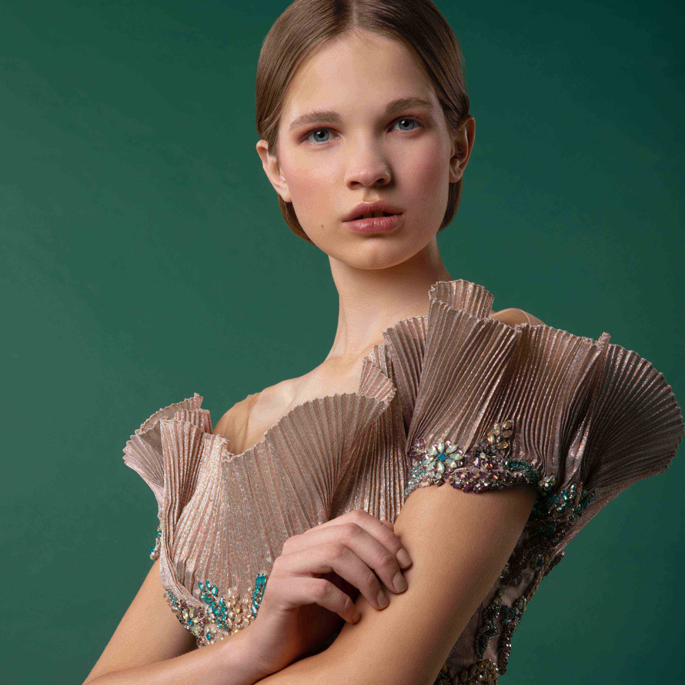 Off shoulder garment with bejeweled bodice over a structural high-low pleated gown.